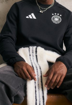 Personne portant un sweat-shirt noir de football d'Allemagne avec logo et symbole Adidas, tenant une écharpe blanche duveteuse Adidas avec des rayures noires.