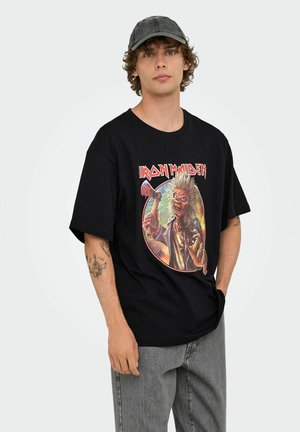 Zwarte katoenen T-shirt met een kleurrafische afbeelding van een personage dat een bijl vasthoudt, met de tekst "Iron Maiden" erboven. Model draagt een grijze jeans en een pet.