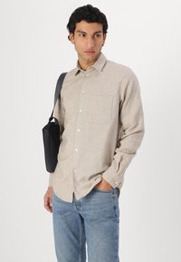 Cămașă butonată de culoare bej deschis, cu buzunar, mâneci lungi și fabricată dintr-un material texturat, asociată cu blugi din denim albastru și o geantă de umăr neagră.