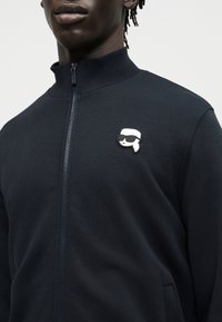 Άνδρας φοράει μαύρο τζάκετ με φερμουάρ, με μια μικρή λευκή καρφίτσα που απεικονίζει ένα στυλιζαρισμένο πρόσωπο με γυαλιά ηλίου στο αριστερό στήθος.