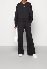 Svart överstort sweatshirt med vit Nike-logga, tillsammans med lösa svarta sweatpants med logga. Vita sneakers och en liten vit väska.