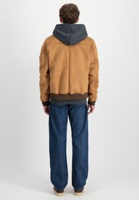 Chaqueta bomber de cuero marrón con capucha gris, puños y dobladillo acanalados, combinada con pantalones de denim azul sueltos y zapatos de color claro.