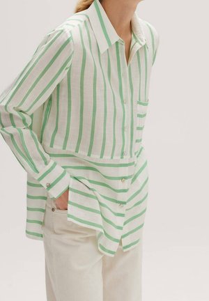 Femme portant une chemise à rayures verticales et horizontales vertes et blanches, boutonnée, à manches longues, et un pantalon beige.
