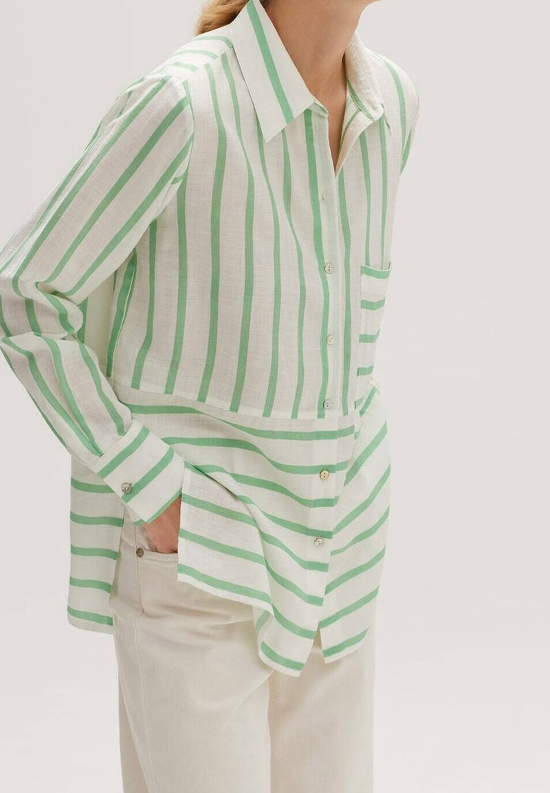 Femme portant une chemise à rayures verticales et horizontales vertes et blanches, boutonnée, à manches longues, et un pantalon beige.