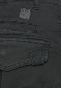 Schwarzer Stoff mit einem aufgenähten Alpha Industries Markenzeichen über einer Klapptasche mit texturiertem Zuglasche.