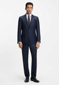 Abito navy con vestibilità su misura, chiusura a un bottone, rever a rotonda e pantaloni con pence. Abbinato a una camicia bianca e una cravatta a fantasia.