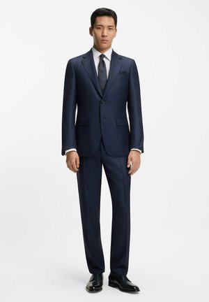 Abito navy con vestibilità su misura, chiusura a un bottone, rever a rotonda e pantaloni con pence. Abbinato a una camicia bianca e una cravatta a fantasia.