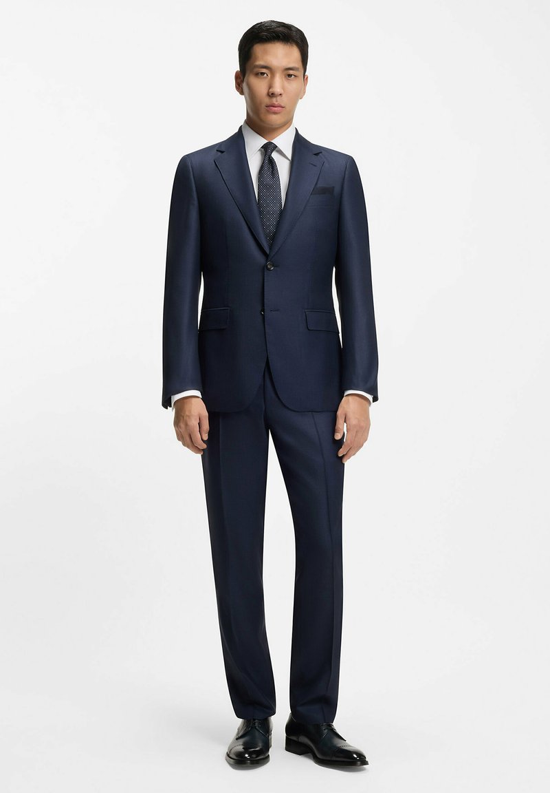 Abito navy con vestibilità su misura, chiusura a un bottone, rever a rotonda e pantaloni con pence. Abbinato a una camicia bianca e una cravatta a fantasia.