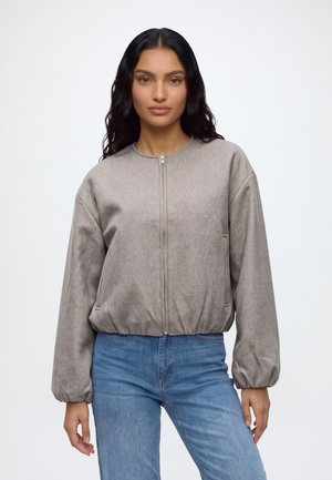 Femme aux longs cheveux foncés portant une veste grise zippée avec un ourlet élastique et un jean bleu, debout devant un fond blanc uni.