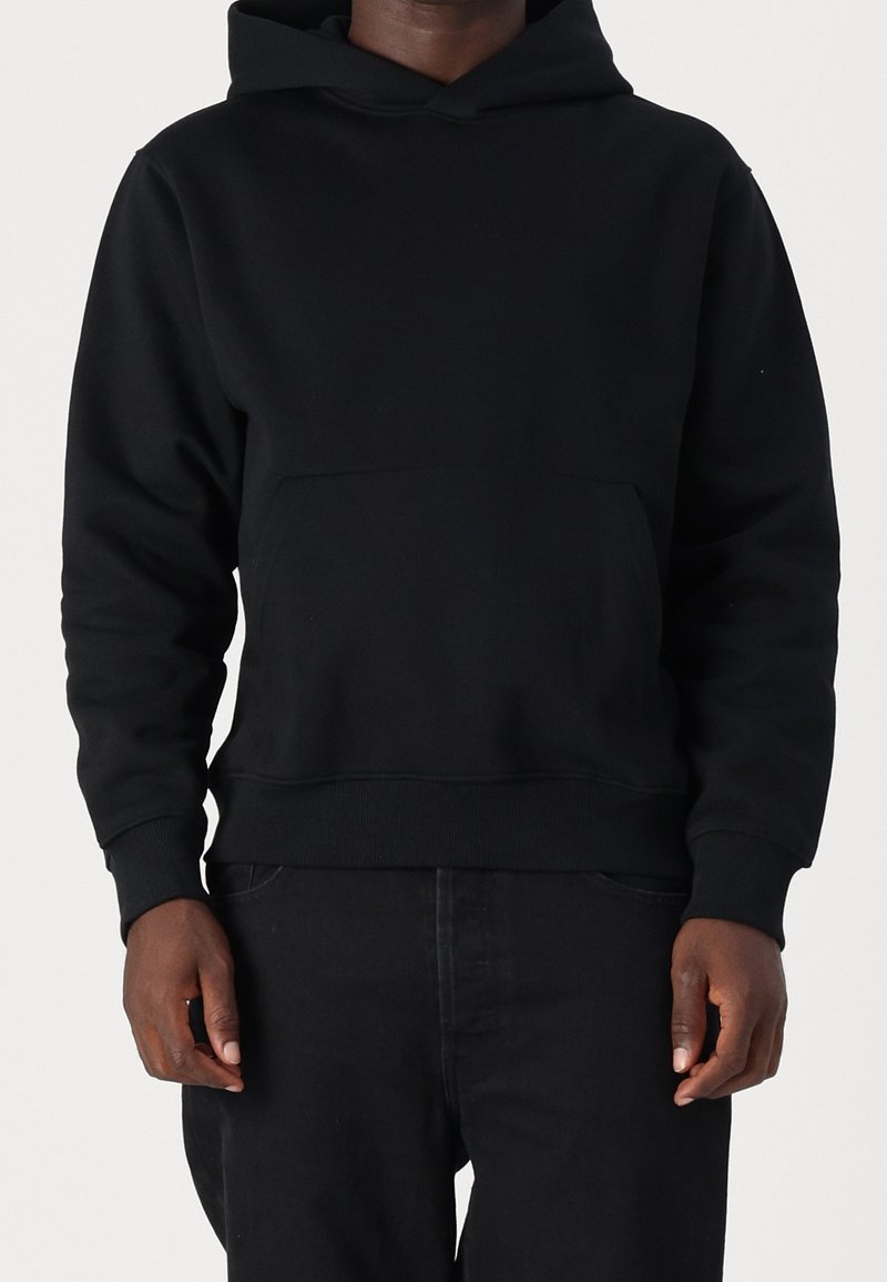 Homme portant un sweat à capuche noir uni avec poche avant et un pantalon noir, debout devant un fond clair uni.