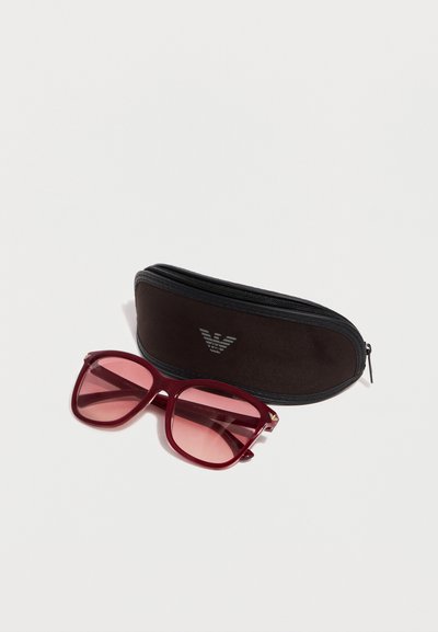 Ορθογώνια γυαλιά ηλίου με κόκκινη απόχρωση και ταίριαστο σκελετό δίπλα σε μαύρη θήκη με φερμουάρ και ασημένιο λογότυπο σε λευκό φόντο.