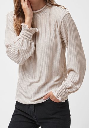 Femme portant un chemisier beige côtelé à manches longues avec des poignets élastiques volantés et un pantalon noir, une main touche son visage, l'autre dans la poche.