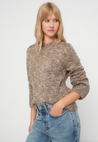 Világosbarna kötött pulóver, texturált mintával, bordás mandzsettákkal és magas nyakkivágással, amelyet magas derekú kék farmernadrággal viselnek.