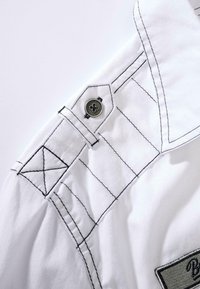 White shirt collar featuring black stitching and a decorative button. Detailed shoulder with an angular design and a logo patch.
