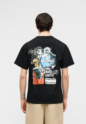 Personne portant un t-shirt noir avec un graphisme coloré, un globe et des textes disant « le monde d'aujourd'hui », « pourquoi pas vous ? » et « Transformez le monde ! »