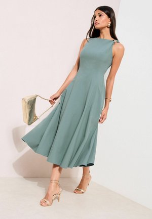 Ein grünes, ärmelloses Midikleid mit ausgestelltem Rock und einer Schulterverzierung. Kombiniert mit goldfarbenen Riemchenhighheels und einer metallisch goldenen Clutch.