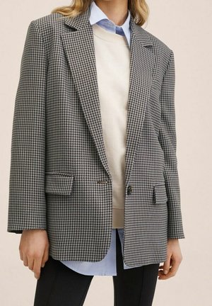 Kvinde iført en grå og sort pied de poule-blazer over en hvid sweater og blåt skjorte, kombineret med sorte bukser mod en lys baggrund.