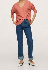 Jasnoróżowy sweter w serek z prążkowanymi mankietami, w połączeniu z niebieskimi, wysokimi dżinsami z rolowanymi mankietami i białymi, szpiczastymi szpilkami.