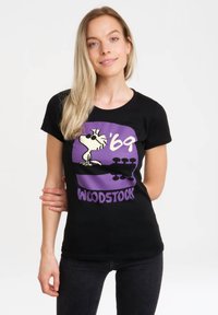 Black cotton t-shirt featuring a graphic of Woodstock in purple, labeled "'69" and "WOODSTOCK" in bold letters. Fitted design.
