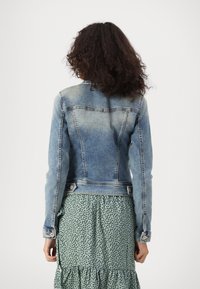 Chaqueta de mezclilla azul claro con un diseño ajustado, textura desgastada, dos bolsillos delanteros y detalles en botones plateados. Llevada sobre un vestido de lunares.