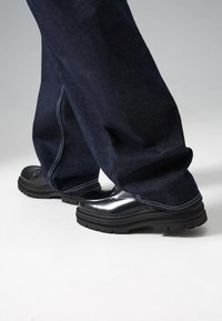 Scarpe in vernice nera con una suola in gomma spessa, abbinate a jeans in denim blu scuro, larghi e con cuciture a contrasto.