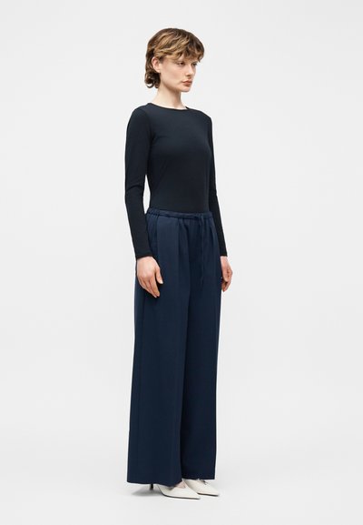 Γυναίκα με κοντά κυματιστά μαλλιά, που φοράει εφαρμοστή μαύρη μπλούζα με μακριά μανίκια, φαρδιά μπλε σκούρα παντελόνια και λευκά τακούνια με μυτερή μύτη, όρθια πλάγια.