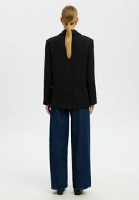 Melnā teksturētā žakete ar izgrieztu apkakli un vienu aizmugurējo šķēlumu, kombinēta ar plašām zila džinsa platām biksēm. Vienkāršs dizains, minimālas deta�ļas.