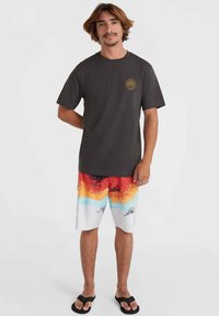 Camiseta de algodón gris oscuro con un logo circular amarillo. Bañador colorido con un patrón degradado en rojo, naranja, azul y blanco. Chanclas incluidas.