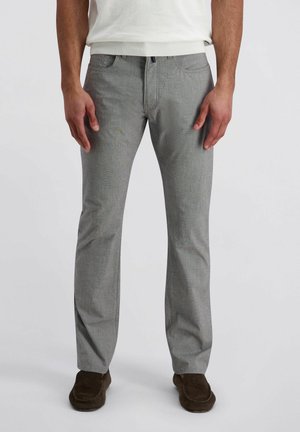 Mann trägt hellgraue, gerade geschnittene Hose, weißes Hemd und dunkelbraune Slipper, steht vor einem schlichten Hintergrund.