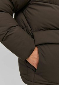 Brun pufferjakke med kviltet design og synlig lomme til hænderne, lavet af et glat, polyester-lignende materiale.