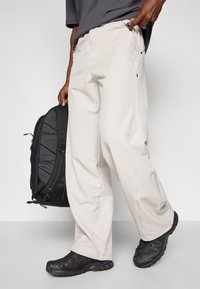 Calças de exterior em bege claro com um corte descontraído, apresentando bolsos e um detalhe de logo. Combinadas com uma mochila preta e sapatos escuros.