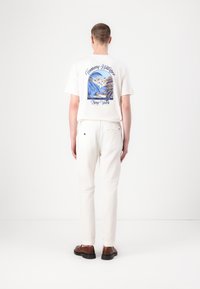Crèmekleurig grafisch T-shirt met een print van een blauw-bruine strandtaferelen op de achterkant, gecombineerd met een witte broek en bruine schoenen.