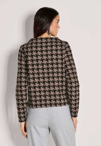 Vrouw met lang donker haar draagt een zwart en beige houndstooth-patroon top en lichtgrijze broek, staand tegen een effen achtergrond.