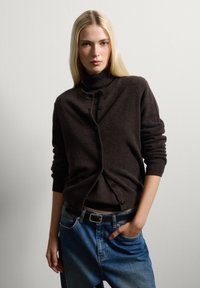 Cardigan in lana marrone con collo alto, caratterizzato da bottoni sul davanti e polsini a coste, abbinato a jeans in denim azzurro chiaro.