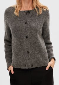 Frau in einem weichen, flauschigen grauen Knöpfen-Cardigan mit langen Ärmeln und schwarzen Hosen, lässig mit den Händen in den Taschen vor einem schlichten Hintergrund.