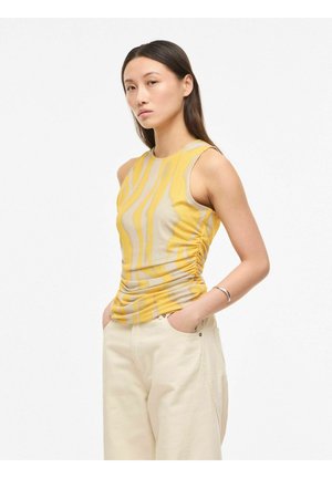 Žena odjevena u žutu i bež prugastu majicu bez rukava s naborima sa strane i kremaste široke hlače, ruke u džepovima, neutralan izraz lica.
