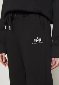 Sorte joggebukser lavet af blødt stof, med elastisk talje og snoretræk samt et hvidt logodesign på venstre lår.