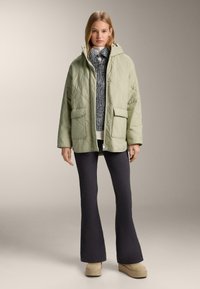 Žena v svetlo zelenej prešívanej bunde, čiernych rozšírených nohaviciach, béžových platformových topánkach a čierno-bielom pletenom svetri stojaca pred jednofarebným pozadím.