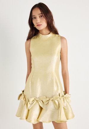 Lesklé zlaté šaty s vysokým výstrihom, bez rukávov a rozšíreným lemom, zdobené veľkými mašľami na spodnej strane.