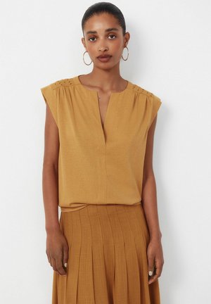 Femme portant un haut jaune moutarde à manches courtes avec une encolure en V et une jupe plissée assortie, debout devant un fond blanc.