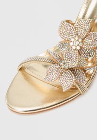 Kuldne kontsaga sandaal lillekujundusega, kaunistatud mitmevärviliste litritega. Sile metallik tekstuur ja avatud varbaga stiil.