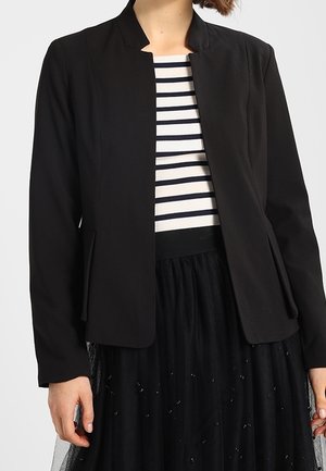 Femme portant un blazer noir cintré sur un haut rayé blanc et noir et une jupe en tulle noire avec des embellissements subtils.