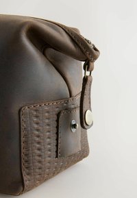 Punguță din piele maro cu un accent țesut texturat, dotată cu fermoar argintiu și închidere cu capsa. Prezintă o suprafață moale și plisată.