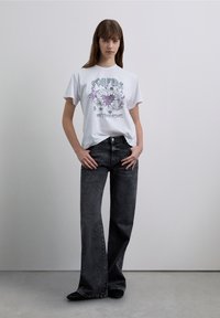 Wit grafisch T-shirt met paarse en zwarte opdruk, gecombineerd met zwart, hoogtailleren uitgewerkte jeans. Model staat tegen een neutrale achtergrond.
