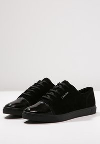 Nízké černé semišové tenisky s lesklými černými špičkami, černými šňůrkami a logem Calvin Klein po straně každé boty.