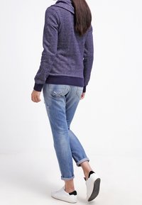 Sudadera de textura púrpura con cuello alto, combinada con jeans claros azul claro con dobladillos y zapatillas blancas con detalles en negro.