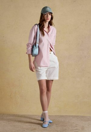 Noor naine kannab roosa triibulise särgi, valgete lühikeste pükste, sinist mütsi, sinist õlakotti, läbipaistvaid valgeid sokke ja helesiniseid kontsakingi, seisab beeži seina taustal.