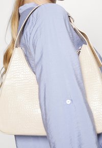 Beige kabelka z umělé kůže s krokodýlím vzorem, trojúhelníkového tvaru, se zipovým uzávěrem, držena proti světle modré texturované blůze.