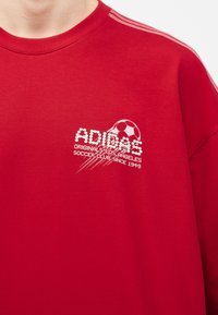 Κόκκινο φούτερ με λευκό λογότυπο Adidas, γραφικό με ποδοσφαιρική μπάλα και κείμενο «Originals Los Angeles Soccer Club Since 1949» στο στήθος.