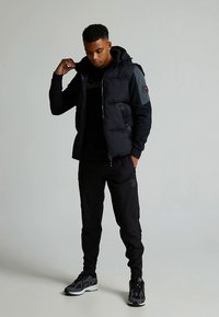 Svart puffjakke med hette, med grå ermer, kombinert med svarte joggebukser og svarte joggesko. Jevn tekstur og tettsittende design.